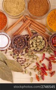 gorgeous setting with cooking spices and herbs (bay leaves, chili powder, coriander, cloves, cardamom pods, cinnamon sticks, garam masala, piri piri, salt, turmeric) on a wooden mat