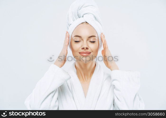 Gorgeous spa woman stands with closed eyes, keeps hands on towel wrapped on head, dressed in white bath robe, has healthy skin, natural makeup, well groomed complexion, poses indoor. Beauty treatment