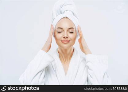 Gorgeous spa woman stands with closed eyes, keeps hands on towel wrapped on head, dressed in white bath robe, has healthy skin, natural makeup, well groomed complexion, poses indoor. Beauty treatment