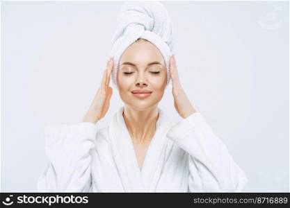 Gorgeous spa woman stands with closed eyes, keeps hands on towel wrapped on head, dressed in white bath robe, has healthy skin, natural makeup, well groomed complexion, poses indoor. Beauty treatment