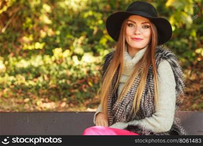 Gorgeous stylish young woman wearing fashionable clothes. Portrait of girl in black hat sitting on bench in park.