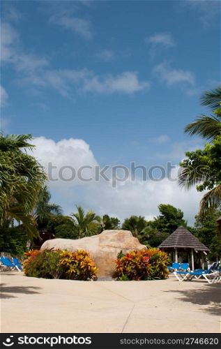 gorgeous swimming pool area with big artificial stone, chairs and bungalow on a tropical resort (surrounded by palm trees)