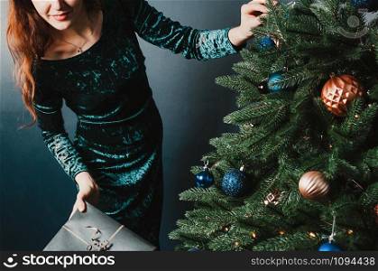 Gorgeous woman in a beautiful dress holding a gift box near Christmas tree, smiling. New Year and Christmas decoration concept. Luxury green, blue, golden colors. Home and family warmth. Banner for holiday theme