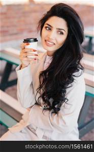 Gorgeous woman with dark hair, eyebrows and well-shaped lips looking aside and smiling drinking cappuccino in takeaway cup sitting at bench outdoors. Beautiful smiling female having nice relaxation