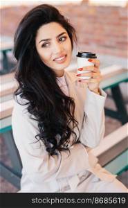 Gorgeous woman with dark hair, eyebrows and well-shaped lips looking aside and smiling drinking cappuccino in takeaway cup sitting at bench outdoors. Beautiful smiling female having nice relaxation