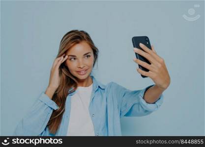 Gorgeous young female holding mobile phone, posing and taking selfie or recording live video for social media while posing in casual outfit against blue background, looking charming and confident. Gorgeous model making selfie on mobile phone