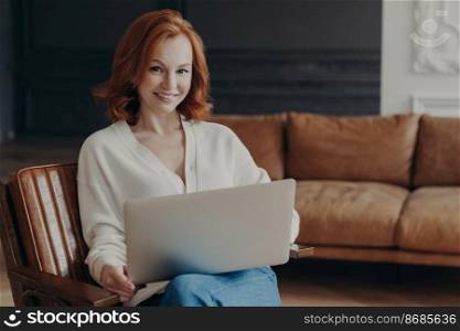 Gorgeous young ginger woman in white jumper sits on armchair with laptop computer, prepares for online interview with new employee, big comfortable sofa in background. Working distantly from home
