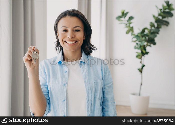 Gorgeous young lady is buying apartment and moving. Satisfied mixed race woman is holding house keychain in her hand near window of living room. Mortgage loan and investment concept.. Gorgeous young lady is buying apartment and moving. Satisfied woman is holding house keychain.