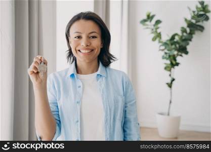 Gorgeous young lady is buying apartment and moving. Satisfied mixed race woman is holding house keychain in her hand near window of living room. Mortgage loan and investment concept.. Gorgeous young lady is buying apartment and moving. Satisfied woman is holding house keychain.