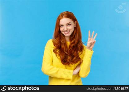 Gorgeous young redhead girl 20s in yellow sweater, smiling carefree making order, reservetion for three people, showing number third with fingers, standing blue background. Copy space. Gorgeous young redhead girl 20s in yellow sweater, smiling carefree making order, reservetion for three people, showing number third with fingers, standing blue background