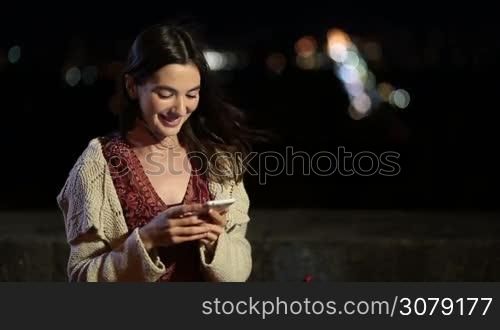 Gorgeous young smiling brunette woman texting message on smartphone at night over colorful citylights bokeh background. Elegant long brown hair female typing on mobile phone while relaxing outdoors in city at night.