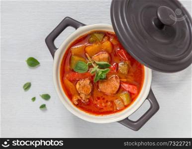 Goulash in a black cocotte with marjoram on white wood.. Goulash in a black cocotte with marjoram on white wood