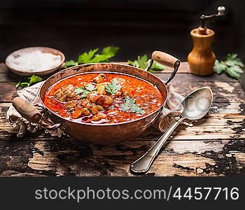 Goulash or stew in copper pan with spoon on rustic kitchen table over dark wooden background