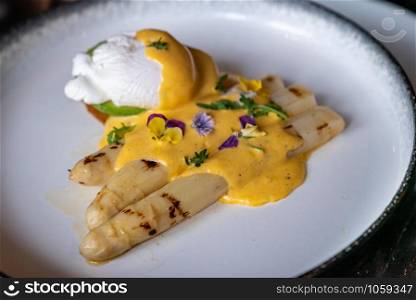 Gourmet grilled white asparagus with poached egg serve with hollandaise savory sauce decorate with edible colorful flowers