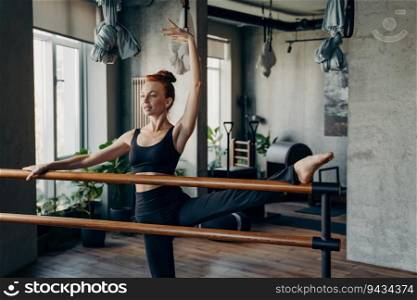 Graceful fit ballerina in black sports bra and leggings stretches with splits at the ballet barre during a Pilates class in the studio. Embracing a healthy and sportive lifestyle.