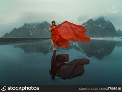 Graceful woman wrapped with red chiffon on beach scenic photography. Picture of person with mountains on background. High quality wallpaper. Photo concept for ads, travel blog, magazine, article. Graceful woman wrapped with red chiffon on beach scenic photography