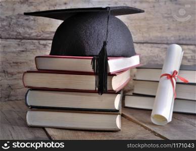 Graduate rolled up in a red ribbon and a black hat on pile of book on wooden background