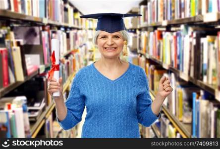 graduation, education and old people concept - happy senior graduate student woman in mortar board with diploma over library background. happy senior graduate student woman with diploma