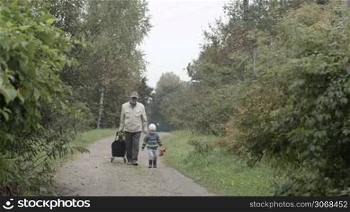Grandad and grandson walking in the countryside. Old man rolling a trolley bag, little boy holdinga toy car.