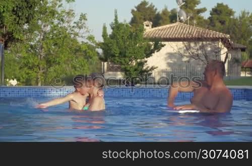 Grandchild and grandparents spending time in outdoor pool at the villa. Grandmother holding boy while he and grandfather playing by splashing water