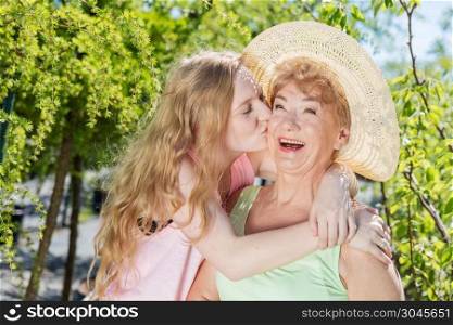 Granddaughter kissing grandmother in a summer garden. Happy family time together. Generations concept. Granddaughter kissing grandmother in a summer garden.
