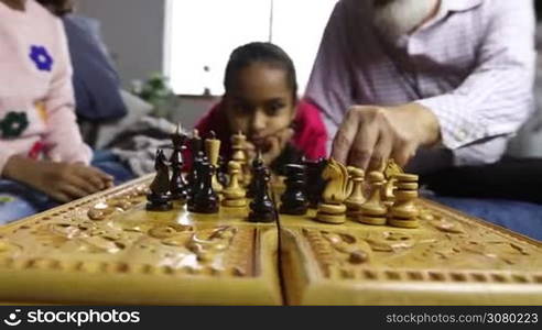 Grandfather and granddaughter playing chess on bed in the living room. Senior man&acute;s and child&acute;s hands moving chess pieces on chessboard during a game with cute small mixed race girl watching the match on background. Closeup. Dolly shot.