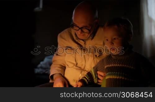 Grandfather and grandson near fireplace at home with fire enlighting their faces. They talking and smiling.