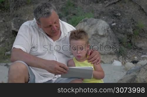 Grandfather and little grandchild spending time outdoor. Boy using table computer while grandpa embracing and talking to him