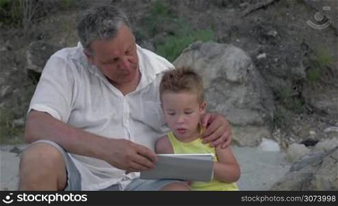 Grandfather and little grandchild spending time outdoor. Boy using table computer while grandpa embracing and talking to him