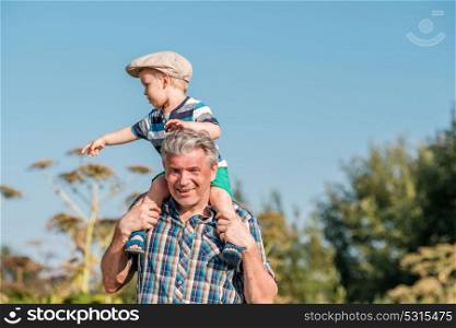 Grandfather carries grandson toddler boy on his shoulders. Child having piggyback ride on his grandfather&rsquo;s back outdoors.