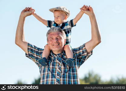 Grandfather carries grandson toddler boy on his shoulders. Child having piggyback ride on his grandfather&rsquo;s back outdoors.