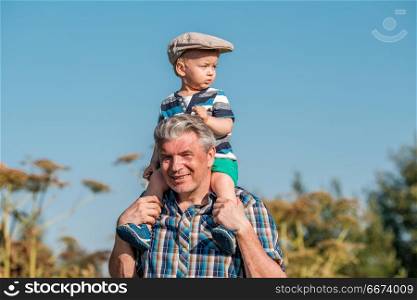 Grandfather carries grandson toddler boy on his shoulders. Grandfather carries grandson toddler boy on his shoulders. Child having piggyback ride on his grandfather&rsquo;s back outdoors.