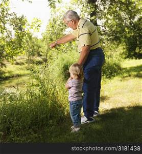 Grandfather holding granddaughter&acute;s hand by creek and pointing.
