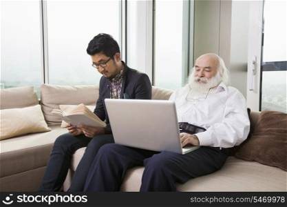 Grandfather using laptop while grandson reading book on sofa at home