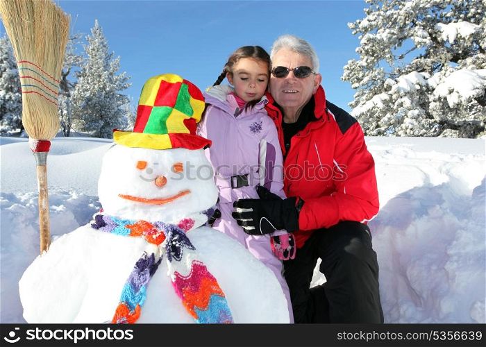 grandfather wearing sunglasses posing with young granddaughter at ski near snowman