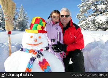 grandfather wearing sunglasses posing with young granddaughter at ski near snowman