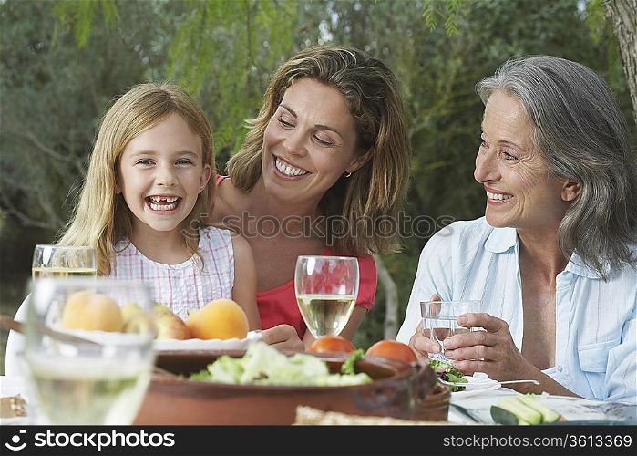 Grandma, mother and daughter (5-6) sitting at table in garden, girl laughing
