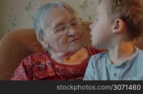 Grandmother and grandson sit on the sofa, grandson sits on grandmother&acute;s knees, then he kisses her