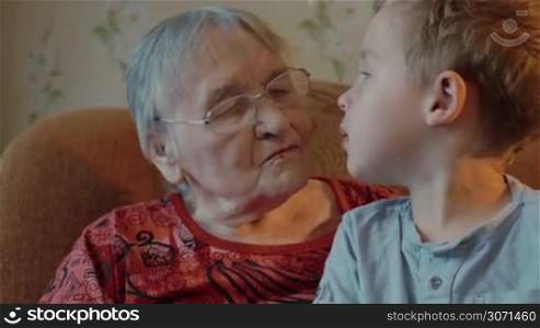 Grandmother and grandson sit on the sofa, grandson sits on grandmother&acute;s knees, then he kisses her