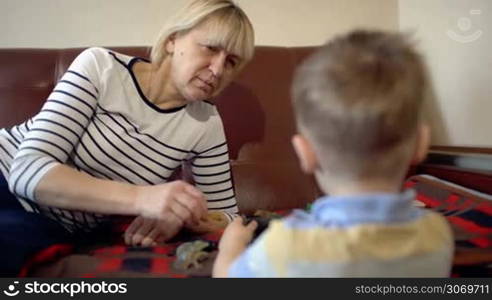 Grandmother and her little grandson playing with toys on the sofa. Boy standing back to the camera, focus on the grandma