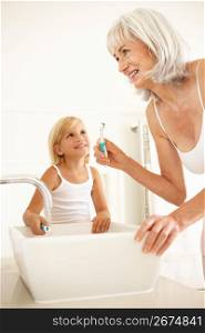 Grandmother Brushing Teeth In Bathroom With Granddaughter Watching