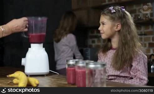 Grandmother pouring fresh red berry smoothie into mason jars from blender shaker jug. Smiling adorable little girl putting drinking straws into mason jars with tasty berry smoothie over domestic kitchen background.
