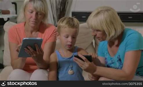 Grandmothers and grandson spending time at home. One grandma using tablet computer and another playing with kid on mobile phone