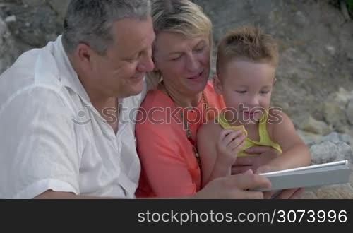 Grandparents and little grandson spending day outdoor. Grandmother and grandfather watching boy playing game on tablet PC