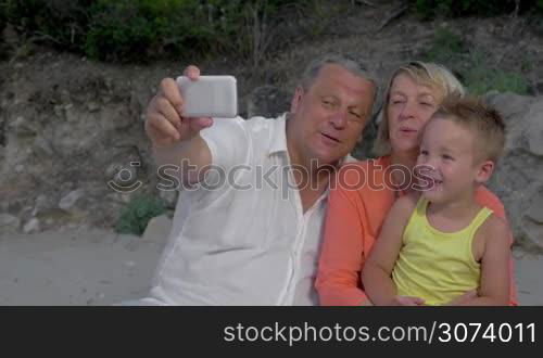 Grandparents are sitting outdoor with their grandson. They are taking selfie shot at smartphone.