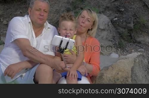 Grandson and grandparents having fun while taking outdoor cellphone selfie using monopod. They making funny faces and laughing