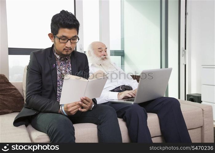 Grandson reading book while grandfather using laptop on sofa at home