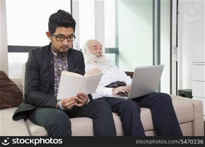 Grandson reading book while grandfather using laptop on sofa at home