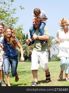 Grandson riding on grandfathers shoulders as family runs in park