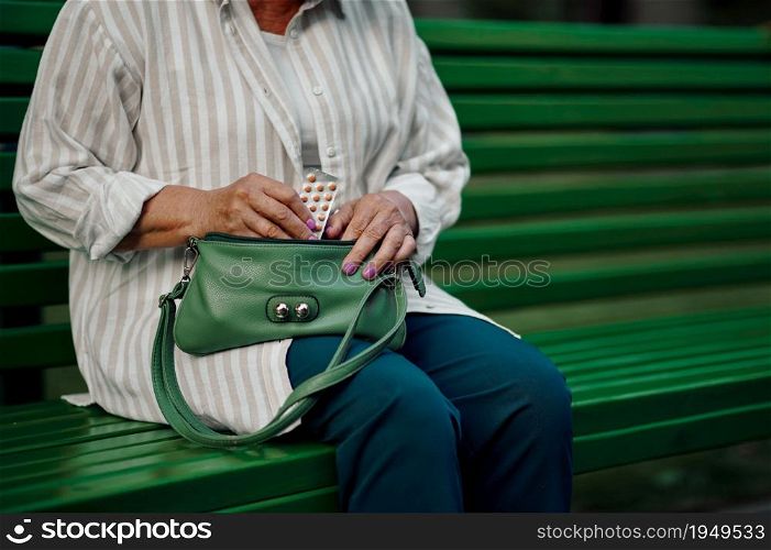 Granny takes the pills out of her bag on the bench in summer park. Aged people lifestyle. Pretty grandmother having fun outdoors, old female person on nature. Granny takes pills out of her bag in summer park